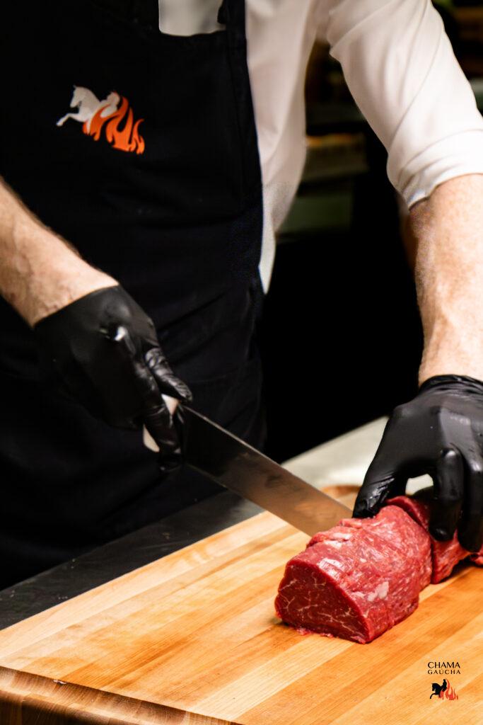 A Gaucho slicing raw filet mignon on a wooden board with a sharp carving knife at Chama Gaucha.